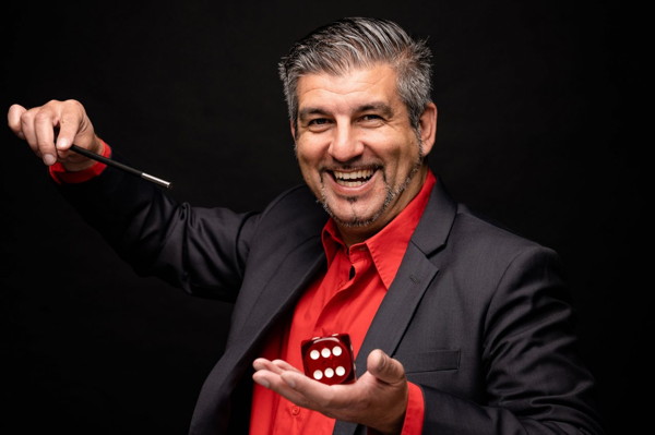 Magier Michele Agusta lächelt und hält in der einen Hand eine Zauberstab und in der anderen einen großen Würfel.